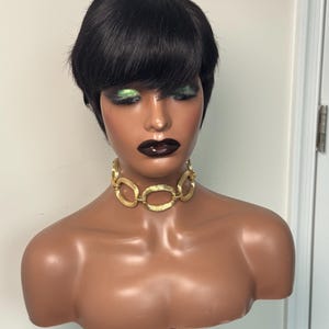 May include: A mannequin head featuring a short, straight black pixie cut wig with bangs. The mannequin has dark skin, green eyeshadow, and dark lipstick. A gold-toned choker necklace with large, textured oval links rests around the neck.