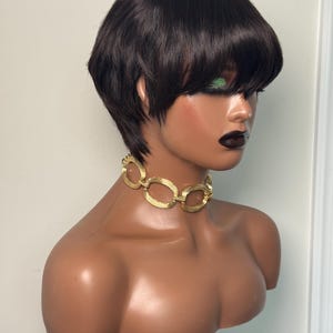 May include: A dark brown short layered wig with bangs on a mannequin head. The mannequin wears a gold-toned choker necklace featuring a series of interconnected oval-shaped links with a textured, ribbed surface finish throughout the metal.