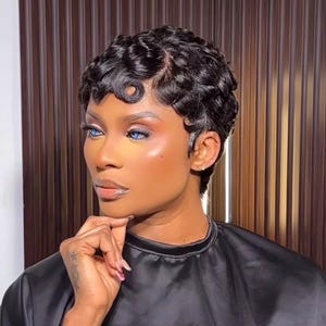 May include: A person with short, dark, wavy hair styled in tight, defined curls. The hair is cropped close to the head with a textured, finger-wave appearance. The individual has bright blue eyes and is wearing a plain black garment.