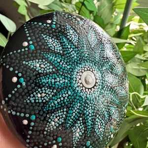 May include: A hand-painted, round stone with a black base, decorated with a teal, white, and silver dot mandala design. The mandala has a central silver accent and is surrounded by small dots. The stone is held against a backdrop of green foliage.