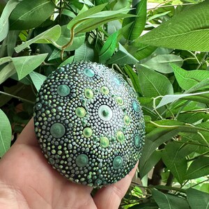 May include: A hand-painted, round decorative stone with a detailed dot pattern in shades of green, teal, and white on a black base. The stone is held against a backdrop of green foliage, highlighting its intricate design and color palette.
