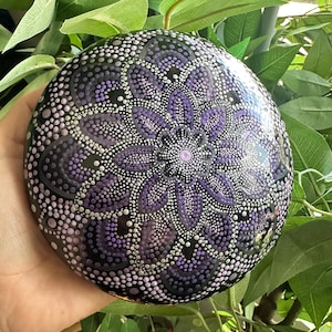 Könnte beinhalten: Ein handbemalter, runder dekorativer Stein mit einem Mandala-Design. Der Stein hat eine schwarze Basis mit einem detaillierten Muster aus lila, lavendelfarbenen und weißen Punkten. Das Design ist zentriert und strahlt nach außen, wodurch ein blütenartiges Aussehen entsteht.