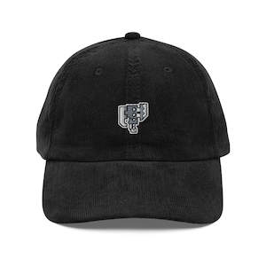 Puede incluir: Gorra de béisbol negra de pana con un pequeño diseño bordado en la parte delantera. La gorra tiene una visera curva y una correa ajustable en la parte trasera. El diseño bordado es un gráfico azul claro y gris.