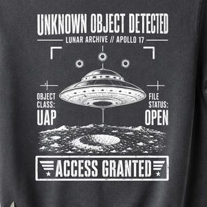 May include: Dark gray t-shirt with a graphic of a UFO over a lunar surface. Text includes "UNKNOWN OBJECT DETECTED," "LUNAR ARCHIVE // APOLLO 17," "UAP," "FILE STATUS: OPEN," and "ACCESS GRANTED." The shirt is hanging on a wooden hanger.
