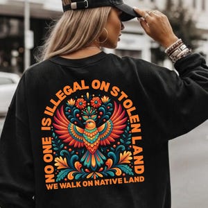 May include: Black sweatshirt with a colorful graphic of a bird and floral design. The text "NO ONE IS ILLEGAL ON STOLEN LAND" is arched above the bird, with "WE WALK ON NATIVE LAND" below. The person is wearing a black cap.