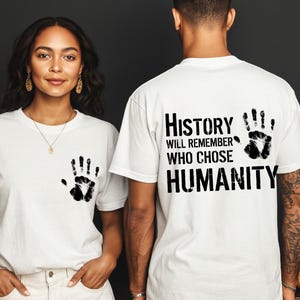 May include: White t-shirts with a black handprint design. One shirt has a handprint on the front, and the other has a handprint and the text "History will remember who chose humanity".