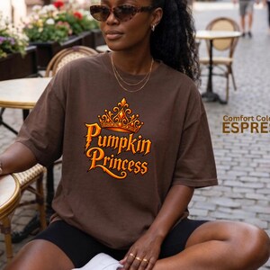 May include: A dark brown short-sleeve t-shirt featuring a graphic of an orange crown above the text Pumpkin Princess in a stylized font. The shirt includes the printed text Comfort Colors 1717 ESPRESSO on the right side of the image.