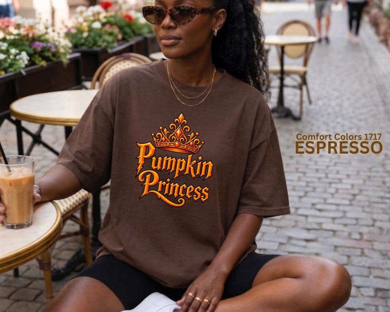 May include: A dark brown short-sleeve t-shirt featuring a graphic of an orange crown above the text Pumpkin Princess in a stylized font. The shirt includes the printed text Comfort Colors 1717 ESPRESSO on the right side of the image.
