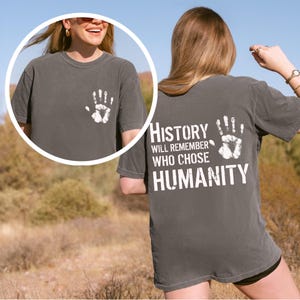 May include: Dark grey t-shirt with a white handprint on the front and back. The back of the shirt features the text "History will remember who chose humanity" in white block letters. The shirt is short-sleeved.