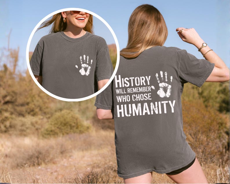 May include: Dark grey t-shirt with a white handprint on the front and back. The back of the shirt features the text "History will remember who chose humanity" in white block letters. The shirt is short-sleeved.