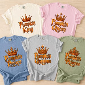 May include: Five short-sleeved crew neck t-shirts in cream, light pink, blue, gray, and sage green. Each shirt features a graphic of an orange crown above text reading Pumpkin King, Pumpkin Queen, Pumpkin Prince, Pumpkin Grandma, or Pumpkin Princess.