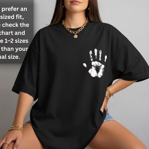 May include: Black t-shirt with a white handprint design on the chest. The shirt has short sleeves and a relaxed fit. Text on the upper left says, "If you prefer an oversized fit, please check the size chart and choose 1-2 sizes larger than your usual size."