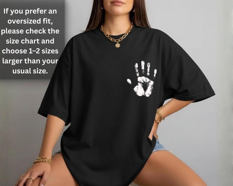 May include: Black t-shirt with a white handprint design on the chest. The shirt has short sleeves and a relaxed fit. Text on the upper left says, "If you prefer an oversized fit, please check the size chart and choose 1-2 sizes larger than your usual size."