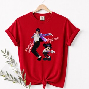 Puede incluir: Camiseta roja con un gr&aacute;fico de Michael Jackson y Mickey Mouse. El dise&ntilde;o muestra a Michael Jackson con sombrero y pantal&oacute;n negros con una chaqueta colorida, y a Mickey Mouse con un atuendo similar. Las palabras "Michael Jackson" y "Mickey Mouse" est&aacute;n escritas en cursiva.