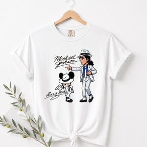 May include: White t-shirt featuring a cartoon illustration of Michael Jackson and Mickey Mouse. Michael Jackson is depicted in a white suit and hat, pointing. Mickey Mouse is also in a white suit and hat. The words "Michael Jackson" and "Mickey Mouse" are written in cursive.