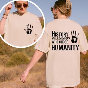May include: Cream-colored t-shirt with a black handprint on the front and the text "History Will Remember Who Chose Humanity" on the back. The shirt is a relaxed fit.