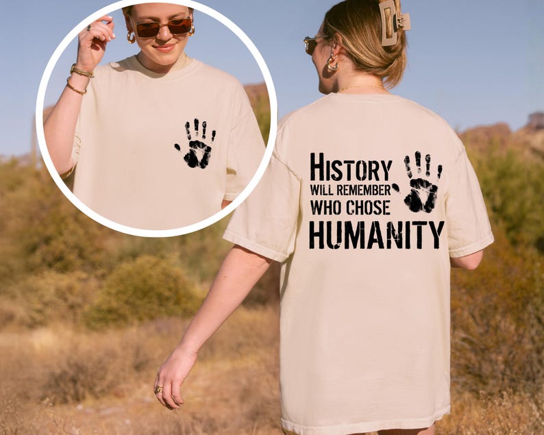 May include: Cream-colored t-shirt with a black handprint on the front and the text "History Will Remember Who Chose Humanity" on the back. The shirt is a relaxed fit.