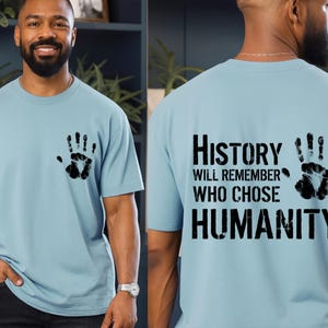May include: Light blue t-shirt with a black handprint on the front and back. The back of the shirt features the text "History Will Remember Who Chose Humanity" in a bold, black font.