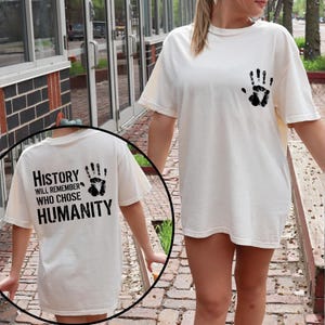 May include: Cream-colored t-shirt with a black handprint on the front and back. The back of the shirt features the text "History will remember who chose humanity" in black block letters.
