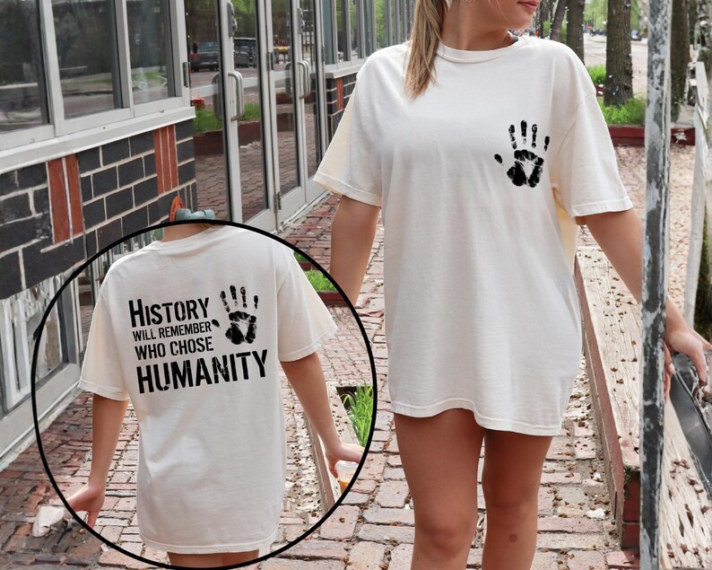 May include: Cream-colored t-shirt with a black handprint on the front and back. The back of the shirt features the text "History will remember who chose humanity" in black block letters.