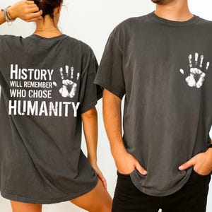 May include: Dark gray t-shirts with white handprint graphics. One shirt has a handprint on the front and the text "HISTORY WILL REMEMBER WHO CHOSE HUMANITY" on the back. The shirts are short-sleeved and have a vintage, worn look.
