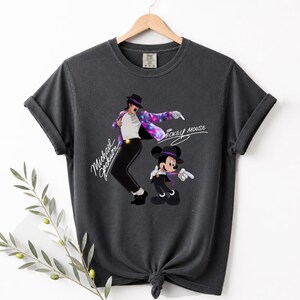 Puede incluir: Camiseta gris oscuro con un dise&ntilde;o de Michael Jackson y Mickey Mouse. El dise&ntilde;o incluye el texto "Michael Jackson" y "Mickey Mouse". La camiseta est&aacute; colgada en una percha de madera. La camiseta es de color gris oscuro.