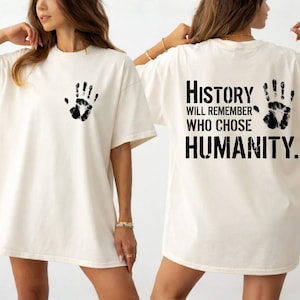 May include: Cream-colored t-shirt with a black handprint on the front and back. The back of the shirt features the text "History will remember who chose humanity."