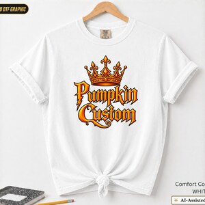 May include: A white short-sleeve t-shirt featuring a central graphic of an orange and gold crown above the text Pumpkin Custom in a stylized orange font. The shirt has a crew neckline and is displayed on a wooden hanger with a knotted hem detail.