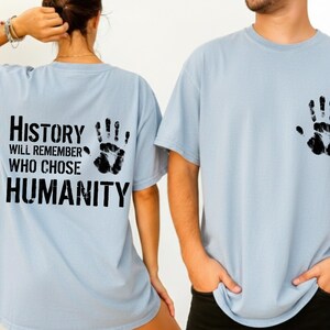May include: Light blue t-shirts with black handprint graphics. One shirt has a handprint on the front and the other has a handprint and the text "History will remember who chose humanity" on the back.