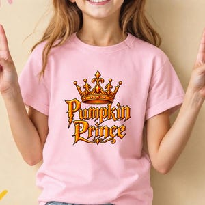 May include: A light pink short-sleeved t-shirt featuring a graphic print of an orange and gold crown positioned above the text Pumpkin Prince in a stylized, bold font with orange and gold gradients and dark outlines on the front center.