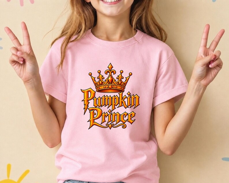 May include: A light pink short-sleeved t-shirt featuring a graphic print of an orange and gold crown positioned above the text Pumpkin Prince in a stylized, bold font with orange and gold gradients and dark outlines on the front center.