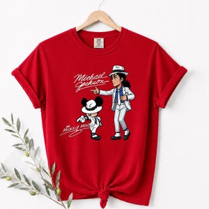 May include: A red t-shirt featuring a cartoon illustration of Michael Jackson and Mickey Mouse in matching white suits and hats. The text "Michael Jackson" and "Mickey Mouse" are written in a cursive font.