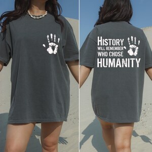 May include: Dark gray t-shirt with a white handprint on the front and back. The back of the shirt features the text "History will remember who chose humanity" in white block letters. The shirt is a casual, oversized fit.