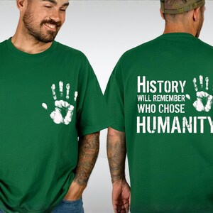 May include: Green t-shirt with a white handprint on the front and the text "History Will Remember Who Chose Humanity" on the back. The shirt is made of a soft material and is suitable for casual wear.