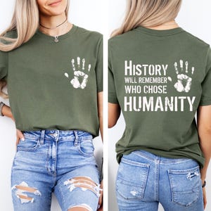 May include: Olive green t-shirt with a white handprint on the front and the text "History will remember who chose humanity" on the back. The shirt is made of a soft material and has a classic crew neck design.