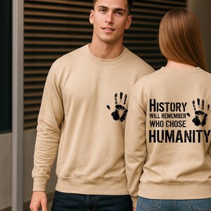 May include: Beige sweatshirts with black handprint designs. One sweatshirt has a handprint on the front pocket, the other has two handprints and the text "History will remember who chose humanity" on the back.