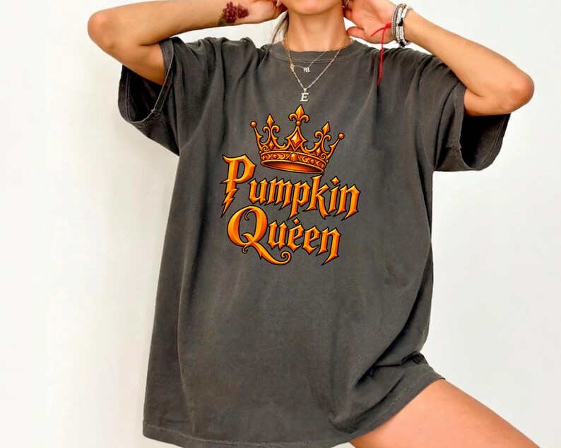 May include: A charcoal gray short-sleeve t-shirt featuring a graphic print on the chest. The design includes an orange crown positioned above the orange text that reads Pumpkin Queen, with a lightning bolt detail integrated into the letter P.