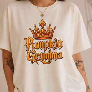 May include: A cream-colored short-sleeve t-shirt featuring a graphic print on the chest. The design includes an orange and yellow crown positioned above the text Pumpkin Grandma, written in a stylized, bold, orange-to-yellow gradient font.