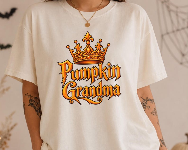 May include: A cream-colored short-sleeve t-shirt featuring a graphic print on the chest. The design includes an orange and yellow crown positioned above the text Pumpkin Grandma, written in a stylized, bold, orange-to-yellow gradient font.