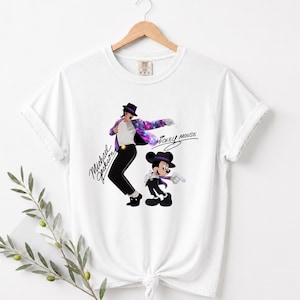 Puede incluir: Camiseta blanca con un gr&aacute;fico de Michael Jackson con una chaqueta con tem&aacute;tica de galaxia y Mickey Mouse. Las palabras "Michael Jackson" y "Mickey Mouse" est&aacute;n escritas en una fuente estilizada. La camiseta est&aacute; colgada de una percha de madera.