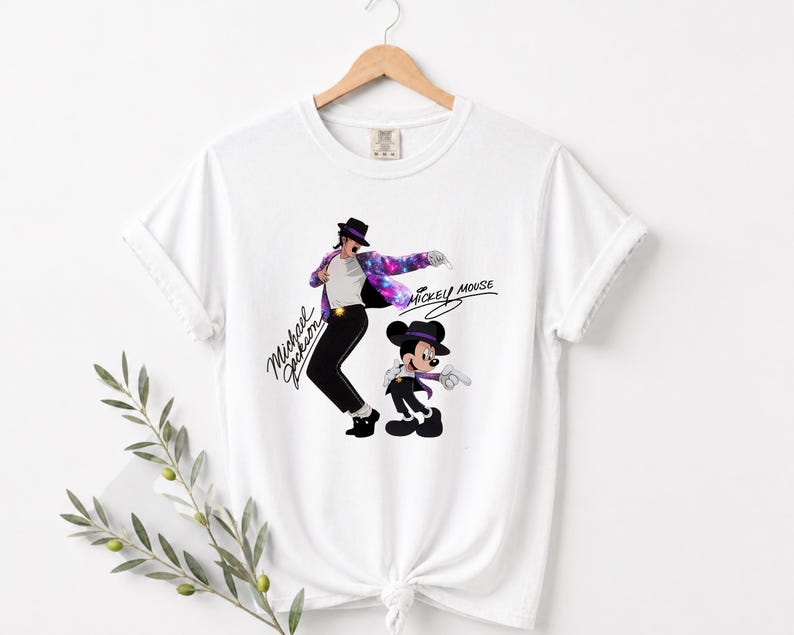 Puede incluir: Camiseta blanca con un gr&aacute;fico de Michael Jackson con una chaqueta con tem&aacute;tica de galaxia y Mickey Mouse. Las palabras "Michael Jackson" y "Mickey Mouse" est&aacute;n escritas en una fuente estilizada. La camiseta est&aacute; colgada de una percha de madera.