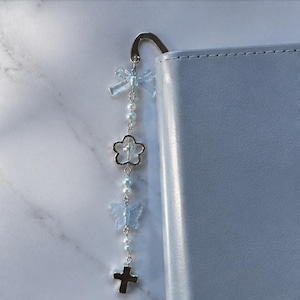 May include: A silver-toned metal hook bookmark attached to a dangling chain featuring light blue translucent plastic bow and butterfly charms, small white spherical beads, a silver-toned open flower charm, and a small silver-toned cross charm.