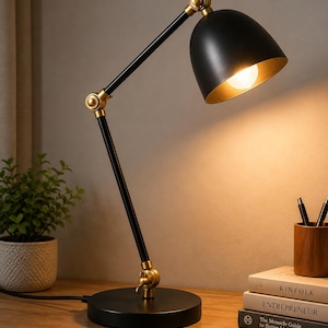 Könnte beinhalten: Eine Schreibtischlampe aus schwarzem Metall und Messing mit verstellbarem Arm und kuppelförmigem Schirm. Die Lampe hat einen runden schwarzen Fuß, zwei Gelenke mit Messingknöpfen und eine warm leuchtende Glühbirne, platziert neben drei Büchern.