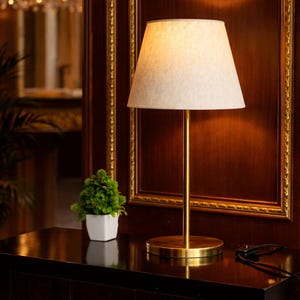 Può includere: Una lampada da tavolo color oro con paralume beige. La lampada ha un gambo sottile e una base circolare. Una piccola pianta in vaso è posizionata nelle vicinanze su una superficie di legno scuro. Lo sfondo presenta un pannello decorativo a parete.
