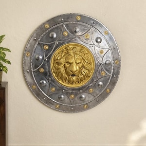 Op de afbeelding: Een cirkelvormig schild met een centraal goudkleurig leeuwenkopreliëf. De buitenrand is afgewerkt in verweerd metallic grijs met gelijkmatig verdeelde zilveren klinknagels en kleine goudkleurige ronde accenten in een geometrisch patroon.