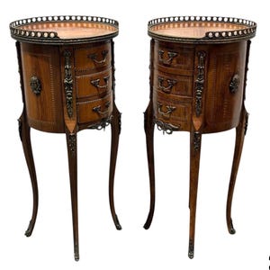 May include: Pair of antique wooden bedside tables. Each table features a round top with a decorative metal rim, three drawers, and ornate metal accents. The tables have slender, curved legs and are finished in a warm brown tone.