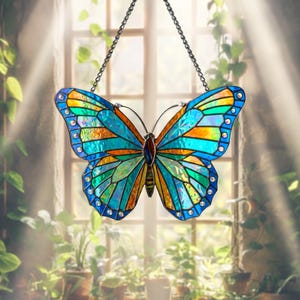 Puede incluir: Un atrapasoles de mariposa de vidrieras, colgado de una cadena. La mariposa presenta un diseño vibrante con piezas de vidrio azul, turquesa, naranja y verde. Los bordes están adornados con pequeñas joyas transparentes. El fondo muestra una ventana y plantas.