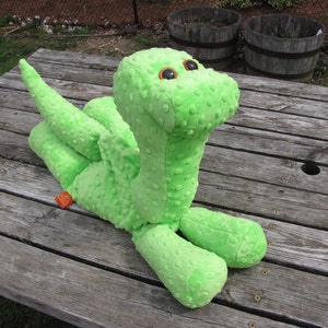 May include: A bright green plush dinosaur toy with large, black eyes. The dinosaur is lying on a wooden surface.