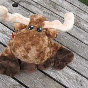 May include: A brown and white plush moose toy with large brown eyes. The moose is sitting on a wooden surface.