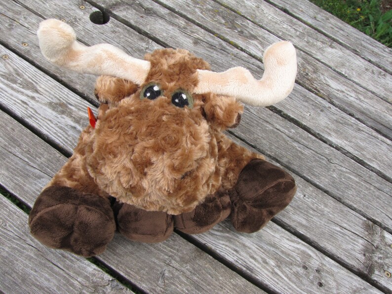 May include: A brown and white plush moose toy with large brown eyes. The moose is sitting on a wooden surface.